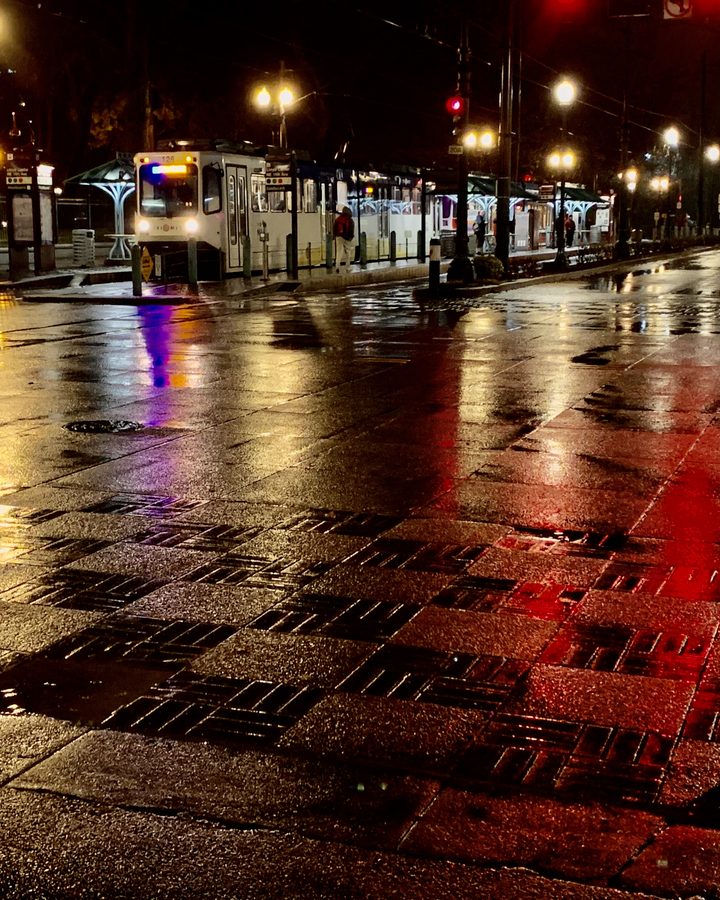 Rain-soaked downtown Portland street at night, streetcar lights reflected in red and gold