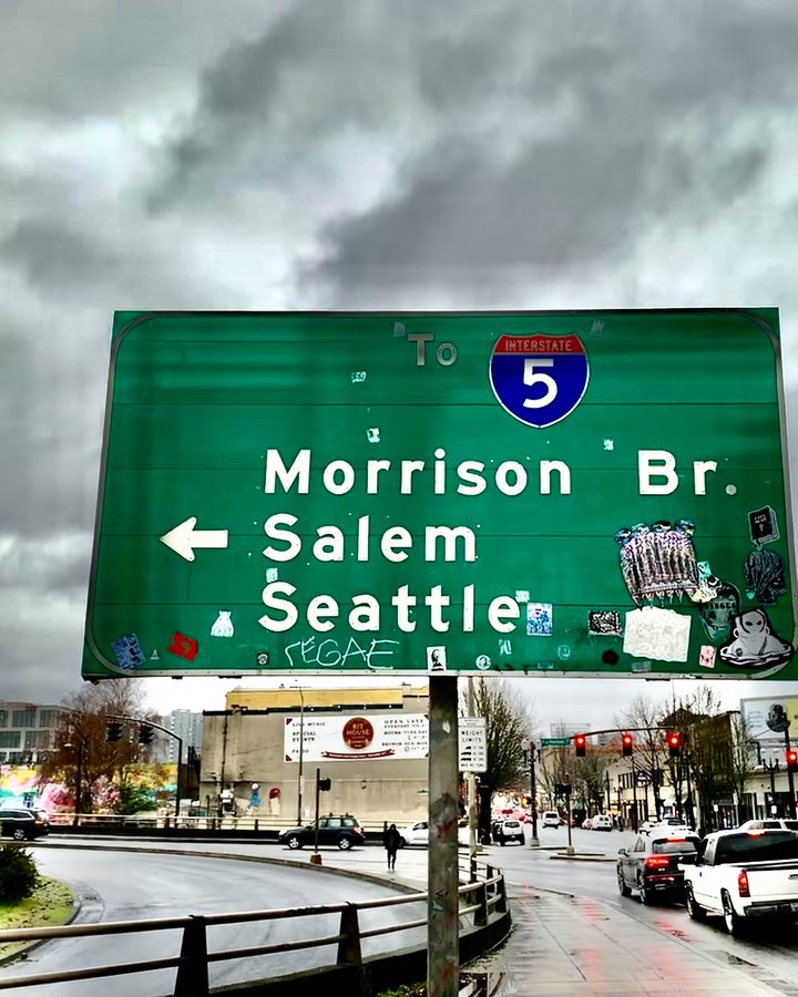 Graffiti-tagged I-5 sign pointing to the Morrison Bridge, Salem and Seattle under storm clouds